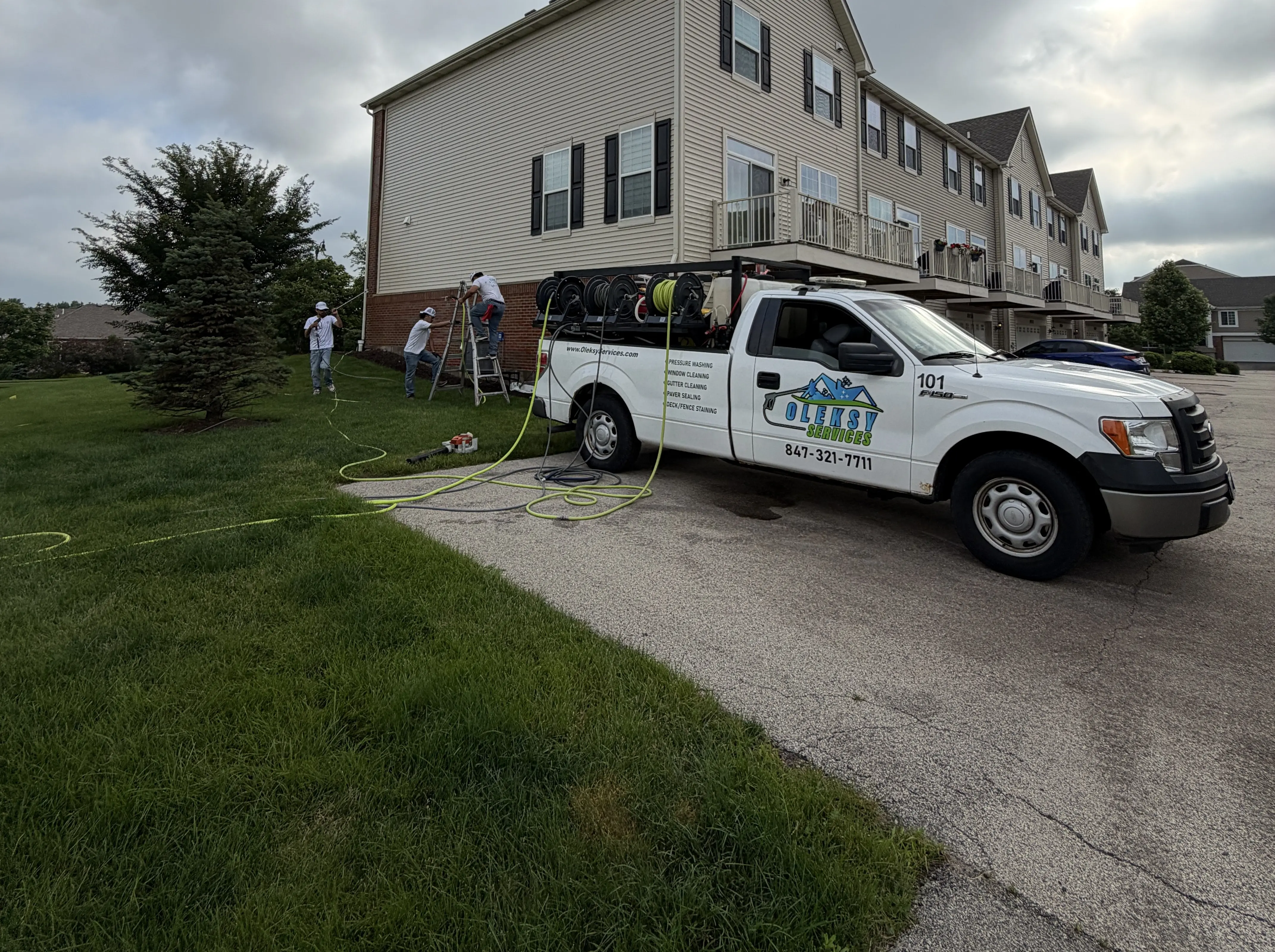 Two Oleksy Services technicians on ladders soft-washing townhome siding with the company truck parked alongside