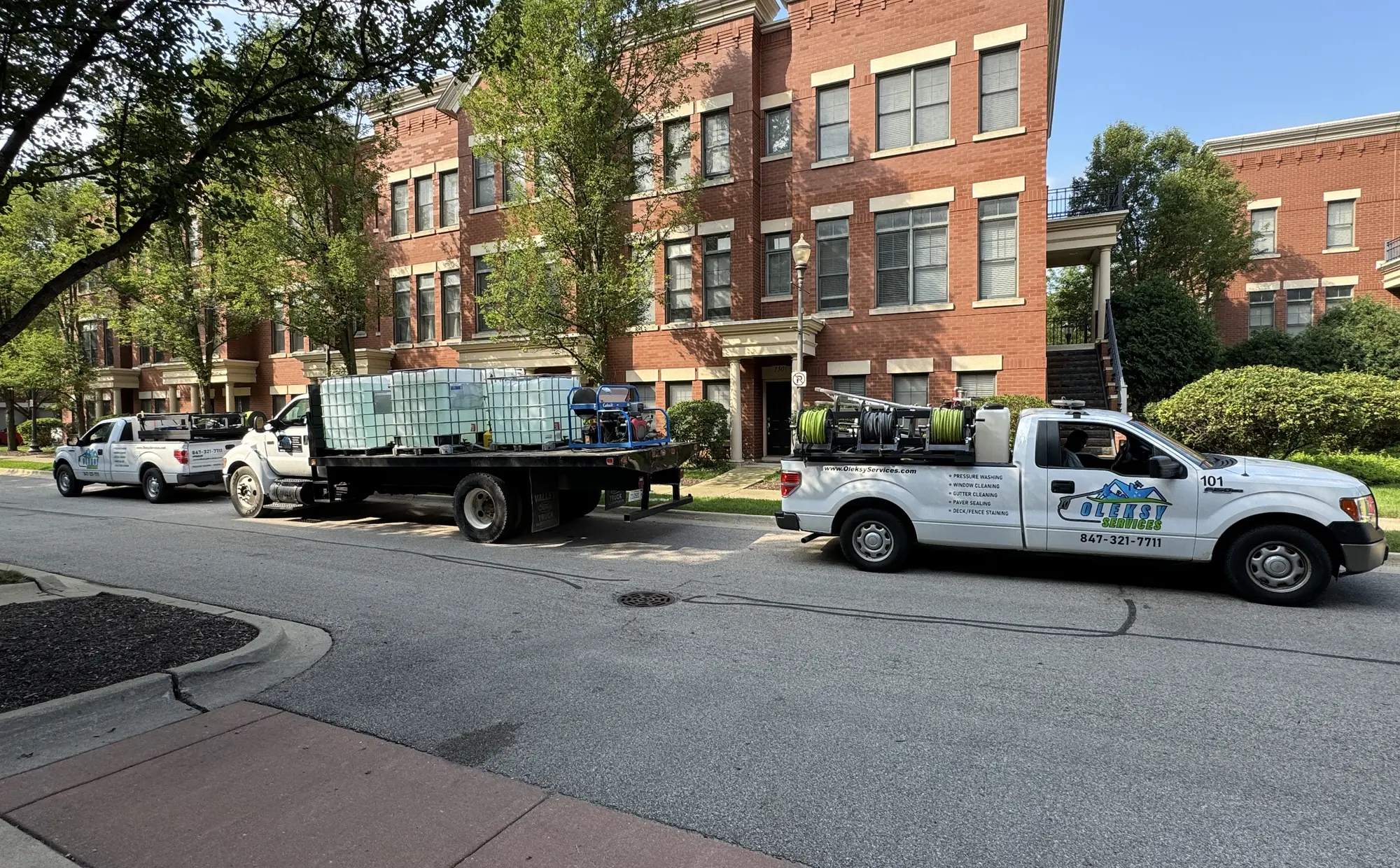 Three Oleksy Services trucks, including a flatbed with water tanks, parked outside a brick condominium building