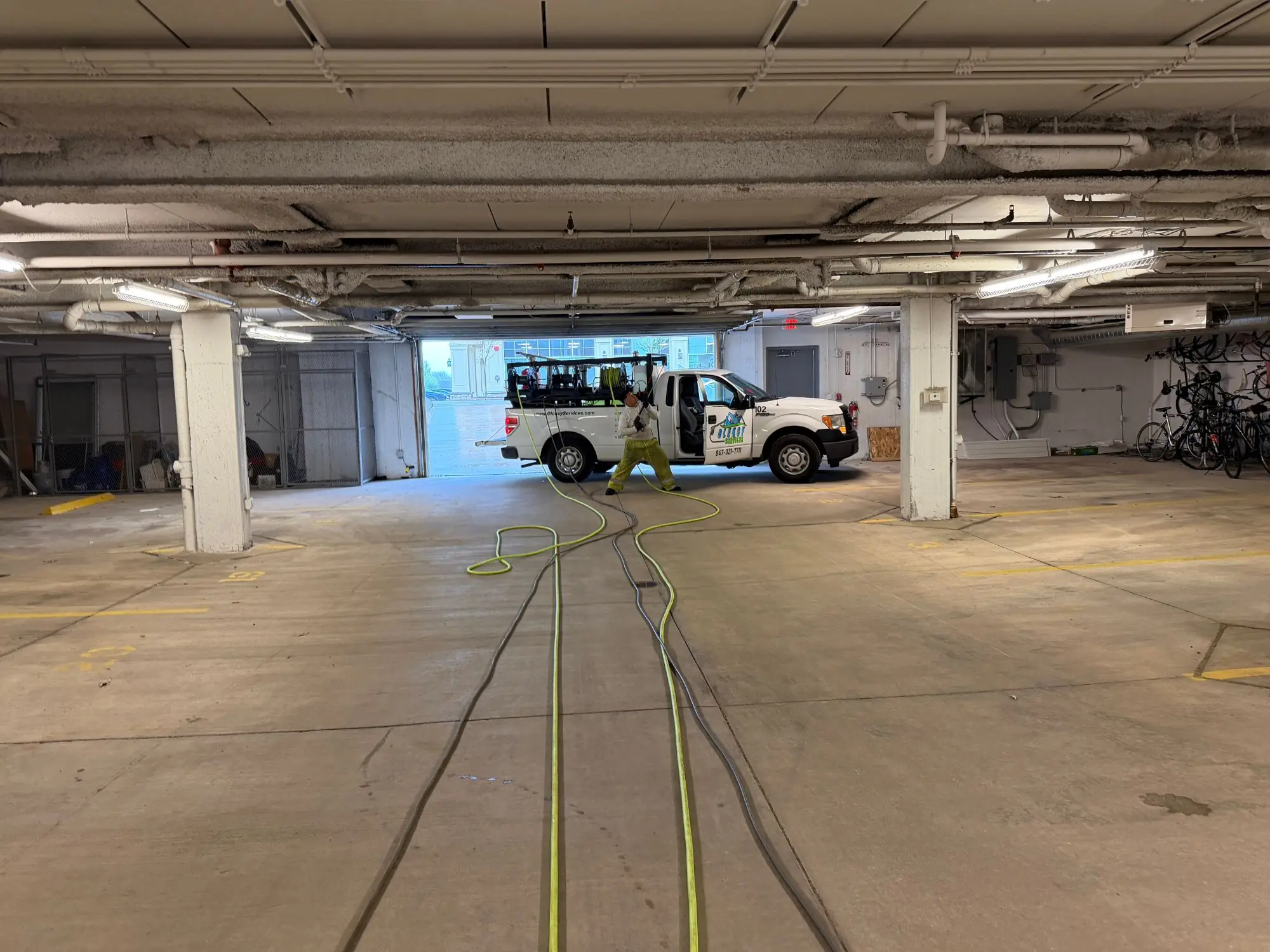 Technician pressure washing the floor of a large underground parking garage, hoses run the full length of the structure